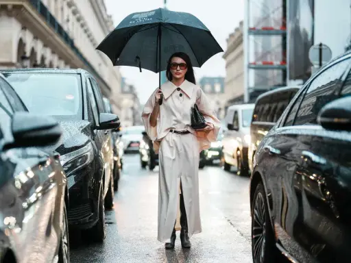 A woman in a chic cream outfit and sunglasses holds an umbrella, showcasing aesthetic rainy day outfits amidst a Parisian street scene with cars.