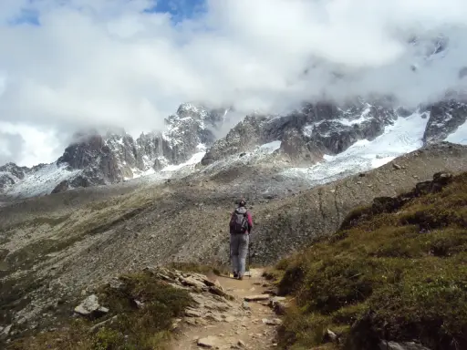 Osoba z plecakiem i kijkami idzie ścieżką w górach. W tle majestatyczne szczyty Alp, częściowo pokryte śniegiem i chmurami. Wspaniały widok na trekking w alpach.