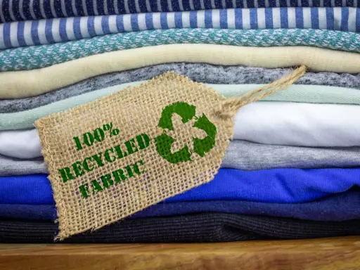 Pile di tessuti ecologici colorati, con un'etichetta che indica "100% Recycled Fabric" e il simbolo del riciclo.