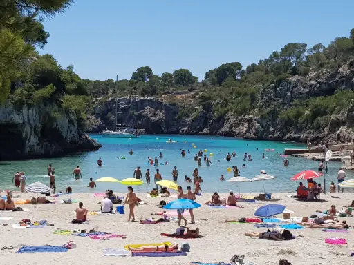 En livlig strand vid Cala Pi, med folk som badar i det turkosa vattnet och solar på sanden under parasoller.