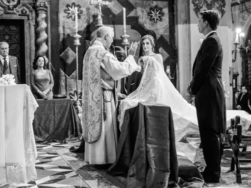 Sacerdote bendice a la pareja durante el rito de la velación. Novia con velo, novio de etiqueta, invitados y altar en iglesia.