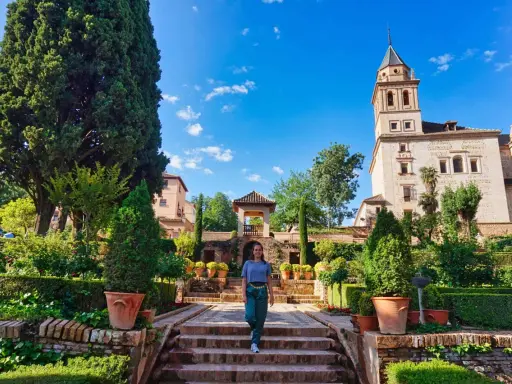 Frau spaziert durch einen üppigen Garten mit Zypressen und einem Turm im Hintergrund. Ein schöner Ort für einen Tagesausflug, vielleicht sogar von Malaga aus.