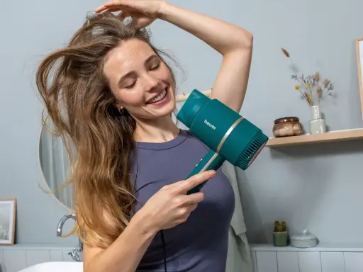 Lächelnde Frau föhnt sich die Haare mit einem türkisfarbenen Föhn. Sie genießt die Pflege ihrer Haare.