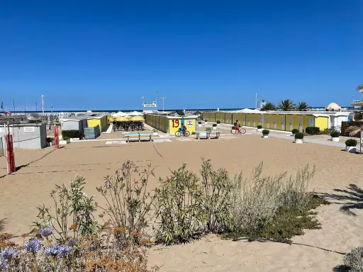 Spiaggia di Riccione con cabine gialle e blu, tavoli da ping pong e bambini in bicicletta al Bagno 19.
