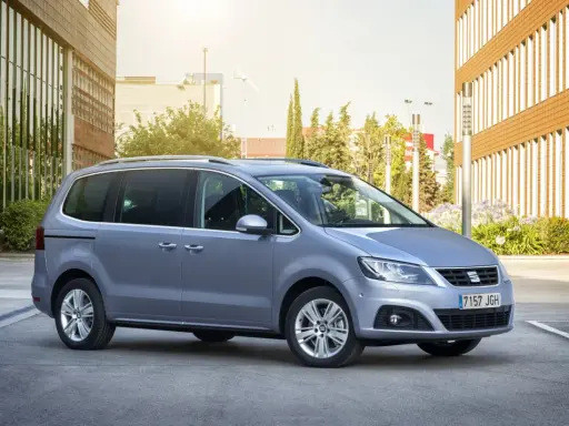 Srebrny Seat Alhambra, rodzinny van, parkuje przed nowoczesnym budynkiem z brązową elewacją.