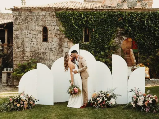 Novios besándose frente a un photocall moderno con arcos blancos y flores. ¡Ideas photocall boda únicas!