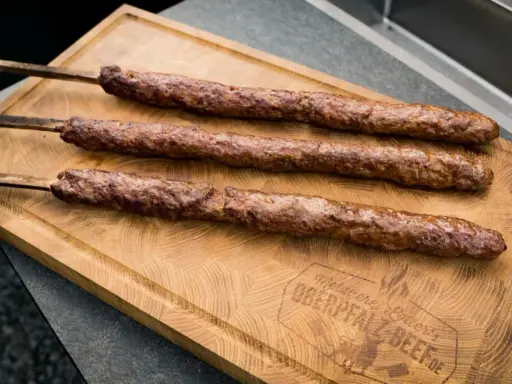 Drei saftige Adana Kebap Spieße liegen auf einem Holzbrett. Perfekt für ein authentisches Adana Kebap Rezept.