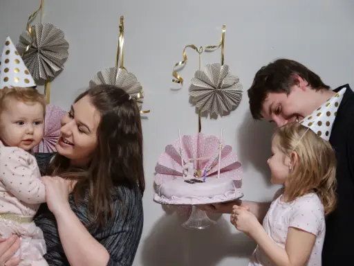 Rodzina świętuje roczek dziecka. Mama trzyma malucha, a tata podaje tort. W tle wiszą papierowe dekoracje, idealne, jak zrobić dekoracje na roczek.