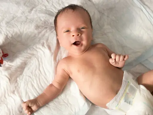 Baby mit offenem Mund, vielleicht im 6-Wochen-Schub, liegt auf einer weißen Unterlage neben einem bunten Spielzeug.