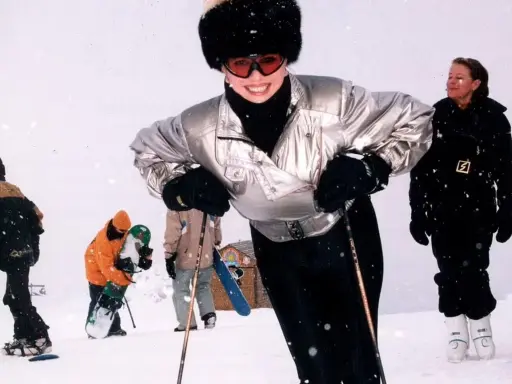 Ivana Trump joven, sonriendo y esquiando con un gorro de piel y gafas de esquí. A su alrededor, otras personas disfrutan de la nieve.