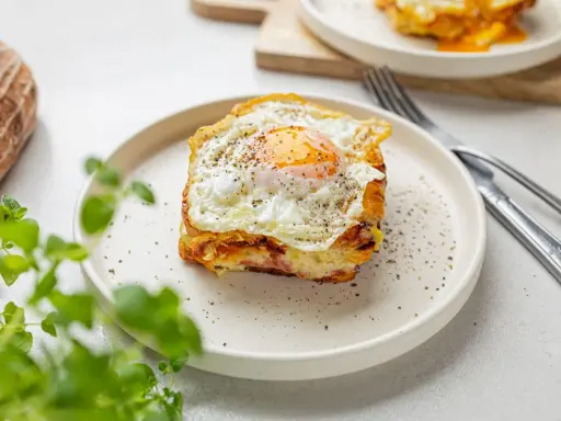 Pyszny croque madame z jajkiem sadzonym na wierzchu, na talerzu, obok chleba i zielonych liści.