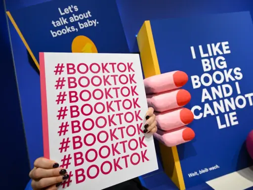 Mani che tengono un cartello con #BOOKTOK ripetuto. Sullo sfondo, un libro blu con la scritta "I LIKE BIG BOOKS AND I CANNOT LIE".