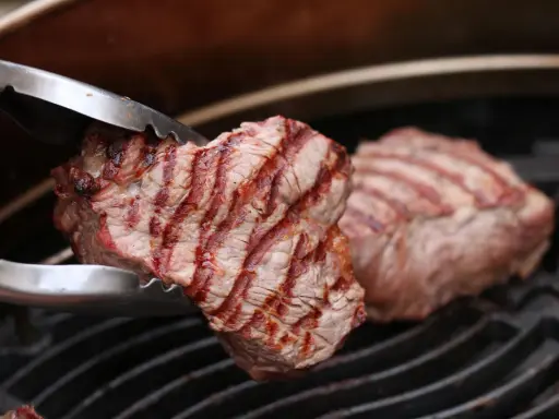 Zangen halten ein saftiges Stück Roastbeef, das auf einem Grill zubereitet wird.