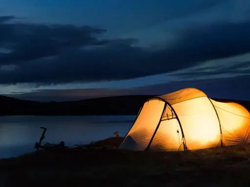 Upplyst tält vid en sjö i skymningen. En cykel står parkerad bredvid. Perfekt för camping Sverige karta-äventyr.