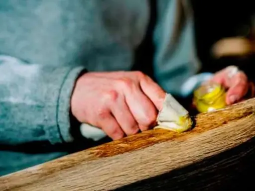 Manos aplicando cera a un trozo de madera, restaurando su belleza. El proceso de decapar madera revela su textura natural.
