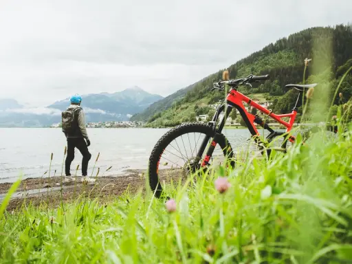En cyklist med hjälm och ryggsäck står vid en sjö med berg i bakgrunden. En mountainbike står redo för äventyr på mtb leder.