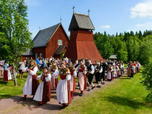 En procession av musiker i folkdräkter, som spelar fiol och gitarr, går förbi en röd träkyrka. Många människor i traditionella kläder, vilka firar midsommar, följer efter.