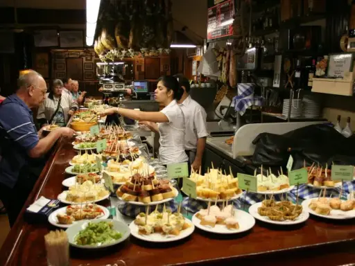 Tapas bar pełen ludzi wybierających przekąski. Na ladzie mnóstwo małych kanapeczek i innych przysmaków.