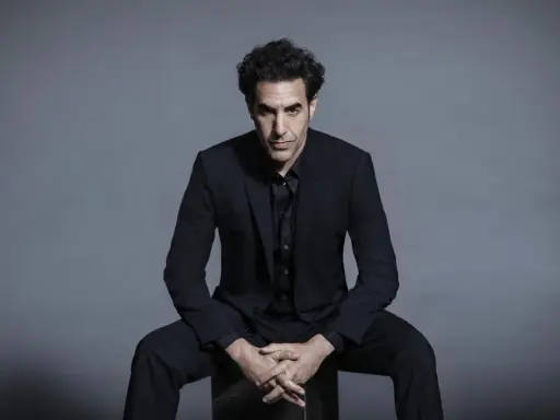 Sacha Baron Cohen, känd för sina kontroversiella filmer, sitter i en mörk kostym med händerna knäppta.