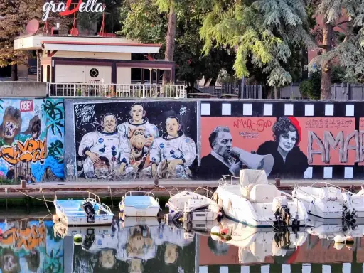 Graffiti colorati sul muro del borgo San Giuliano: astronauti con Alf, personaggi di Amarcord e cani. Barche ormeggiate sull'acqua riflettono i colori.