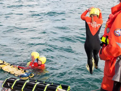 Sauvetage en mer : une sauveteuse en combinaison noire et orange se prépare à plonger. Deux personnes en gilets de sauvetage et casques jaunes flottent près d'une civière. Communication VHF et MMSI essentielle.