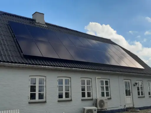 Ett hus med svarta, glänsande nya solceller på taket. Solen reflekteras i panelerna mot en blå himmel med moln.