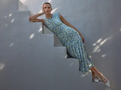 A woman in a floral dress and white heels, embodying a chic formal dress code for women, poses on white stairs.