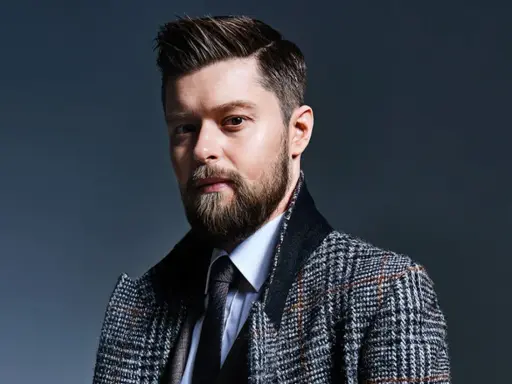 Mężczyzna z brodą i starannie ułożonymi włosami, w eleganckim płaszczu w kratę. Remigiusz Mróz wiek nie jest widoczny, ale styl sugeruje dojrzałość.