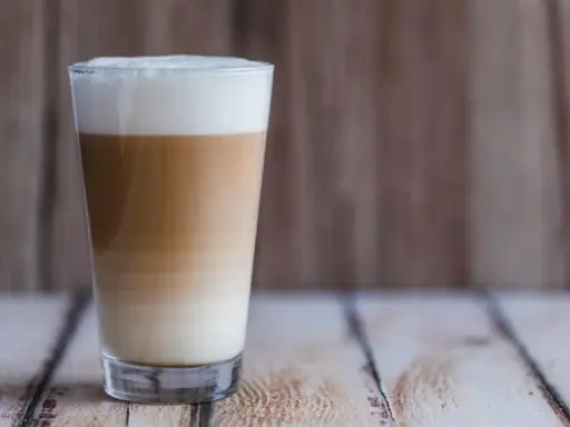 Ein hoher Latte Macchiato mit Mandelmilch, geschichtet in drei Farben, steht auf einem Holztisch.