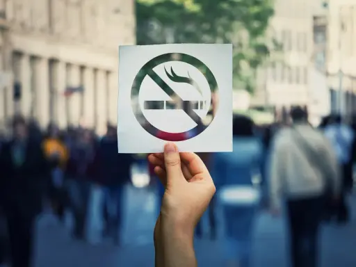 Dłoń trzyma kartkę z symbolem zakazu palenia papierosów na tle tłumu ludzi.