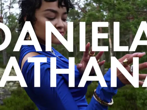 Daniela Rathana i blå klänning i skogen, med texten "DANIELA RATHANA" över bilden. Hon bär smycken och hennes pappa är troligen stolt.