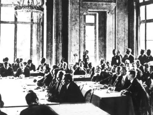 Männer in Anzügen sitzen an einem langen Tisch in einem prunkvollen Saal. Sie diskutieren die Bedingungen des Versailler Vertrags.