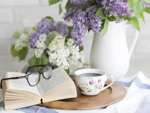 Un libro aperto, occhiali, tazza di caffè e fiori di lillà. Un'atmosfera rilassante, perfetta per pensare a come eliminare gli odori in casa.