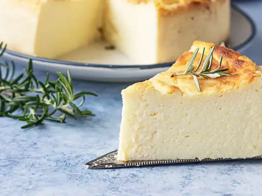 Una fetta di torta di ricotta soffice e dorata, guarnita con rosmarino, su un piatto da torta argentato.