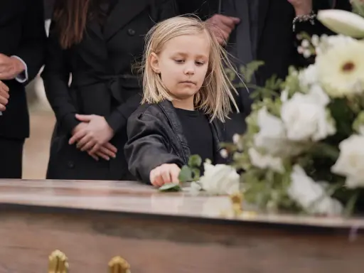 Smutne dziecko na pogrzebie, dotyka kwiatów na trumnie. W tle dorośli w czerni. Czy to dziecko wierzy w przesądy?