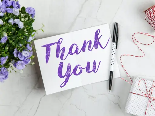 Carte "Thank You" violette, entourée de fleurs bleues, d'un stylo étoilé et d'un cadeau emballé. Une belle façon de remercier quelqu'un pour sa gentillesse.