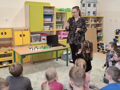 Nauczycielka prowadzi zajęcia kreatywne dla dzieci. Na stole przed nimi leżą klocki Lego, a dzieci z uwagą słuchają.