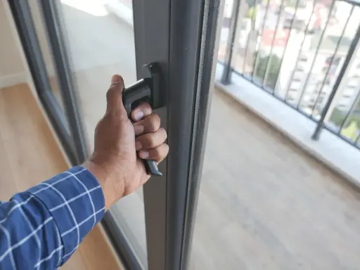 Dłoń chwyta za klamkę, wykonując regulację drzwi balkonowych dwuskrzydłowych. Widok na balkon i miasto.