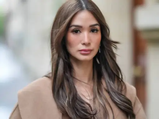 Mujer con cabello castaño largo y ondulado, viste un abrigo beige. Su maquillaje es sutil, un estilo "old money" que realza su belleza natural.