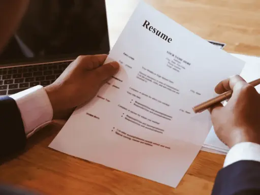 Dłoń trzymająca długopis nad dokumentem CV obok laptopa.