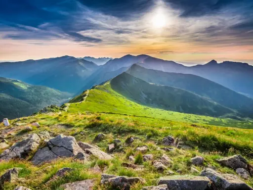 Widok na pasmo górskie o zachodzie słońca. Na pierwszym planie kamienie i trawa, w oddali zielone zbocza i skaliste szczyty. Kasprowy Wierch wysokość robi wrażenie.