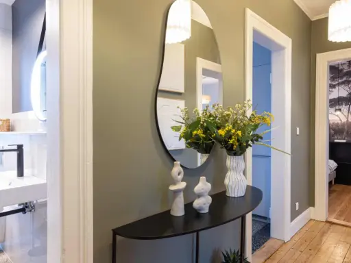 Élégant couloir maison avec miroir asymétrique, vases décoratifs, bouquet de fleurs jaunes et vue sur une salle de bain et une chambre.