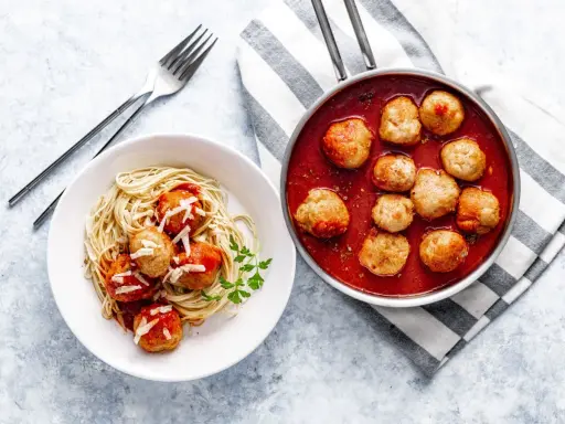 Pyszne pulpety w sosie pomidorowym z makaronem spaghetti, posypane parmezanem. Obok patelnia z gorącymi pulpetami w sosie.