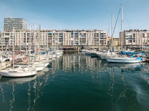Barche a vela ormeggiate nel porto di Tolone, con edifici residenziali sullo sfondo e il cielo azzurro.