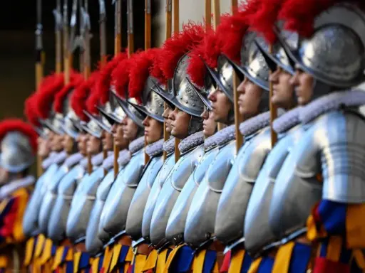 Guardie svizzere in fila, con elmi e piume rosse. Curiosità sulla loro uniforme storica e il loro ruolo.