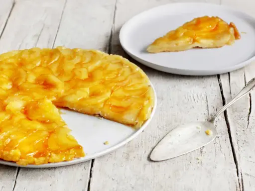 Eine goldbraune Tarte Tatin mit karamellisierten Äpfeln auf einem Teller, daneben ein Stück auf einem weiteren Teller und ein Tortenheber.