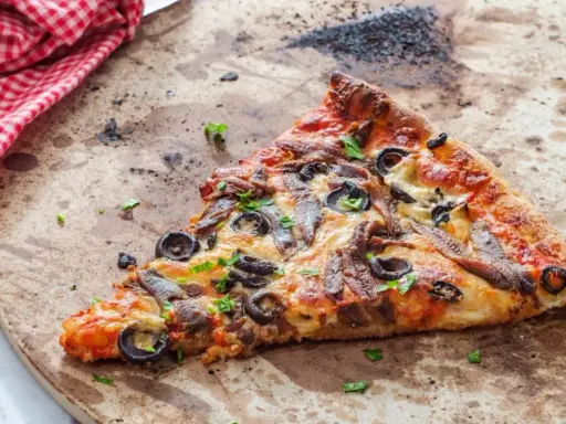 Une part de pizza aux anchois et olives sur une pierre réfractaire, prête pour un nettoyage.
