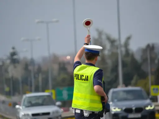 Policjant kieruje ruchem, podnosząc znak stop. W tle widać samochody i słupy oświetleniowe, sugerujące podwyższone **stopnie alarmowe**.