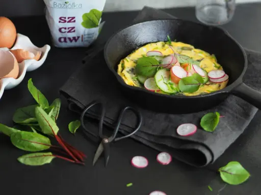 Jak zrobić dobry omlet? Oto inspiracja: puszysty omlet z rzodkiewką, ogórkiem i ziołami na czarnej desce.