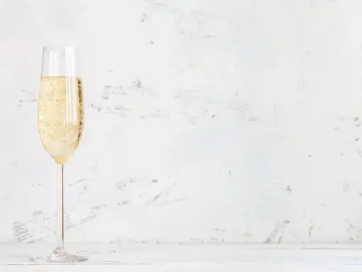 Ett glas champagne med bubblor, kanske en Jaques Selosse, står på ett vitt bord mot en ljus, texturerad bakgrund.